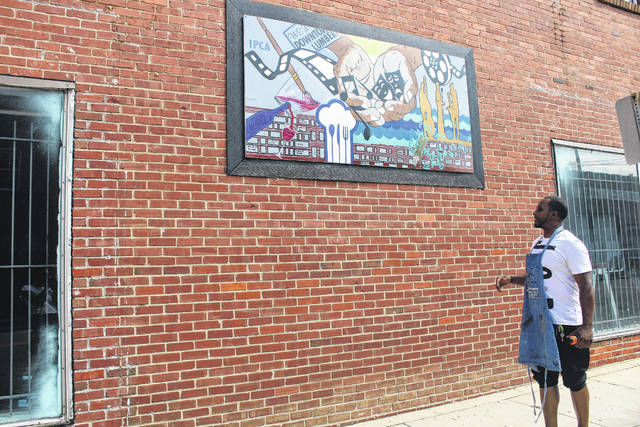 
			
				                                Melvin Morris, founder and curator of the Inner Peace Center for the Arts, stands besides the first of many murals to be featured in The Mural Project. The mural painted by Morris is called “The Offering” and can be viewed at the Third Street wall of the arts gallery located at 302 N. Chestnut St. in Lumberton.
                                 Tomeka Sinclair | The Robesonian

			
		